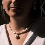 A close-up lifestyle shot of a woman wearing a pearl necklace with a large teardrop-shaped pearl pendant, styled with matching pearl earrings and a white button-down shirt.