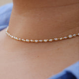 A delicate beaded necklace with small white pearl-like beads and gold spacer beads worn on a person's neck, with a blue shirt visible in the background.
