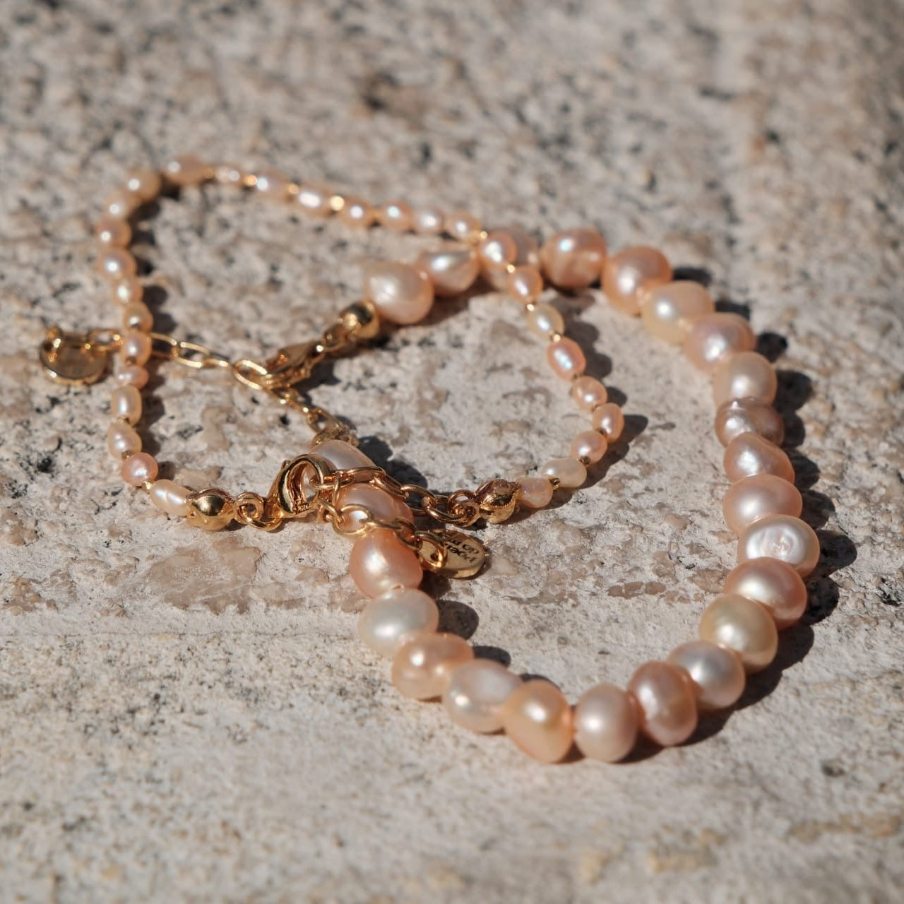 A delicate strand bracelet made of irregular light pink freshwater pearls with gold-toned chain links and clasp, resting on a textured gray stone surface.