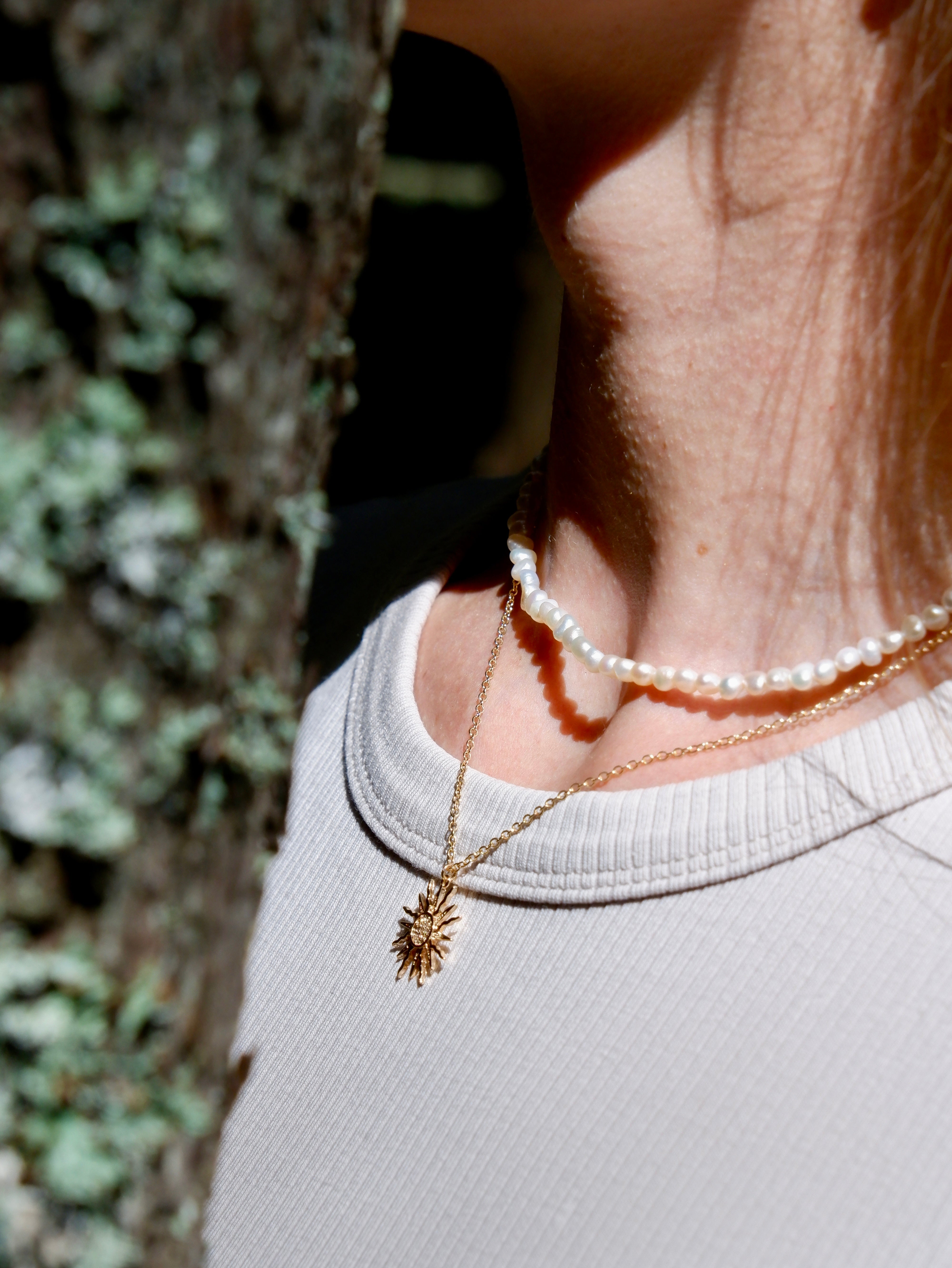 Close-up of a person wearing a gold necklace and pearl bracelet with a blurred natural background