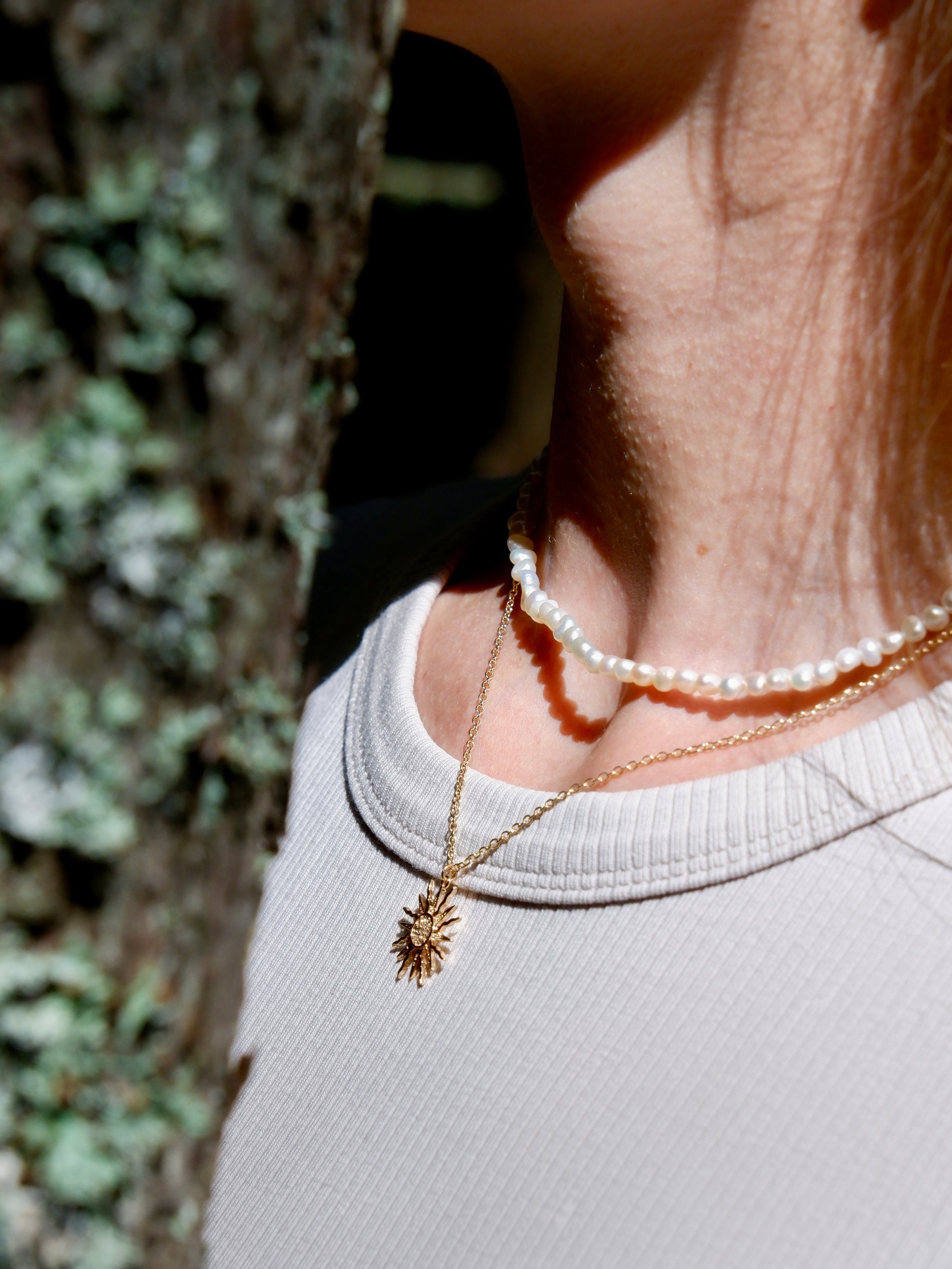 Close-up of a person wearing a gold necklace and pearl bracelet with a blurred natural background
