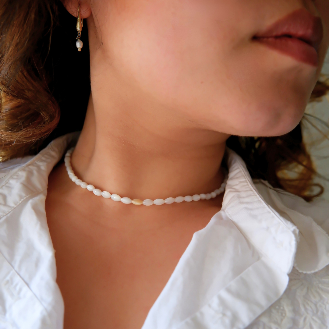 A person wearing a white chocker necklace with rice-shaped mother of pearl beads and gold-plated crimp end beads.