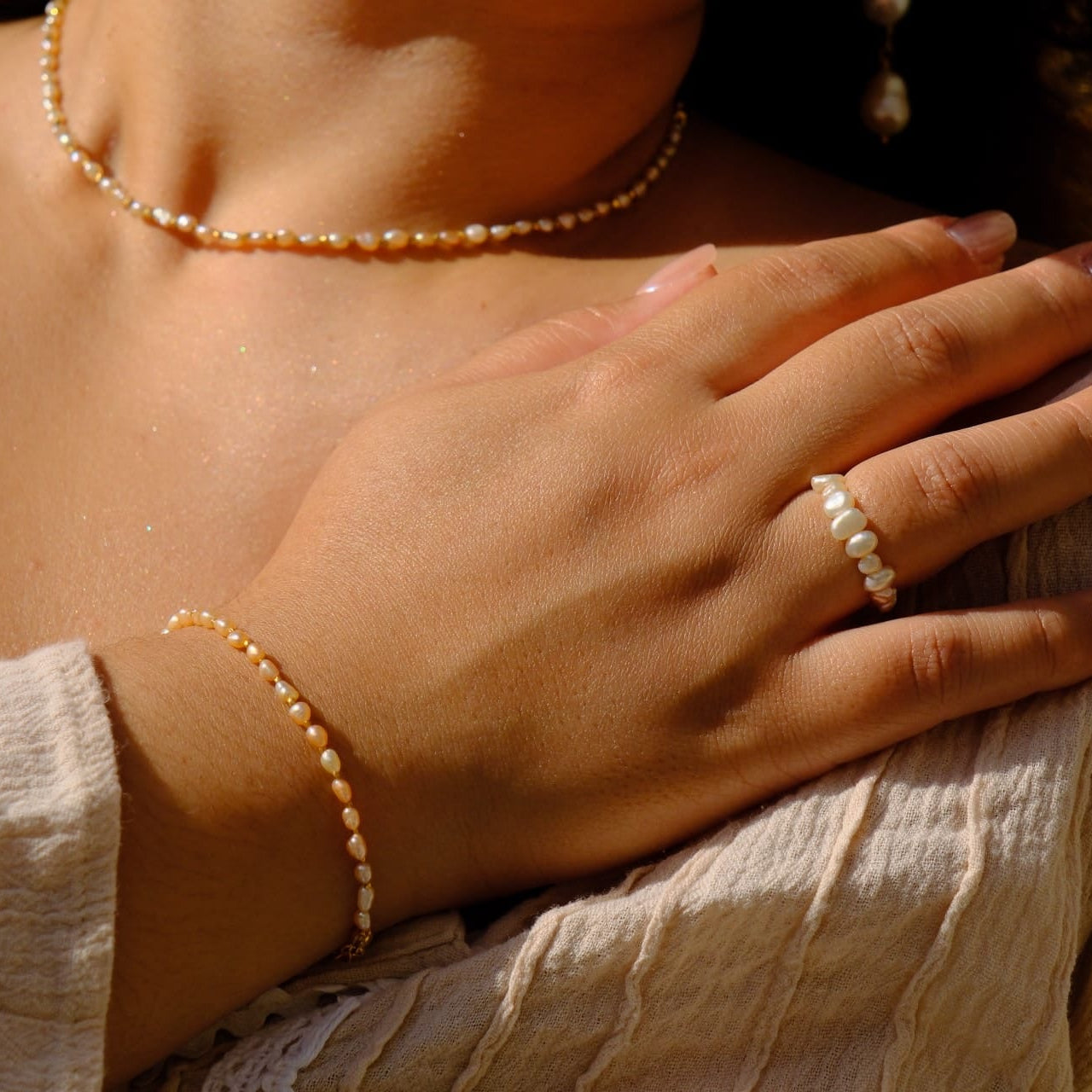 A close-up lifestyle shot of a woman wearing a matching pearl jewelry set, including a pearl necklace, bracelet, and a pearl ring on her finger, styled with a beige textured top.