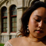 A close-up lifestyle shot of a woman with dark hair wearing a delicate gold chain necklace and pearl drop earrings, set against a blurred background of a classical stone building with arched windows.