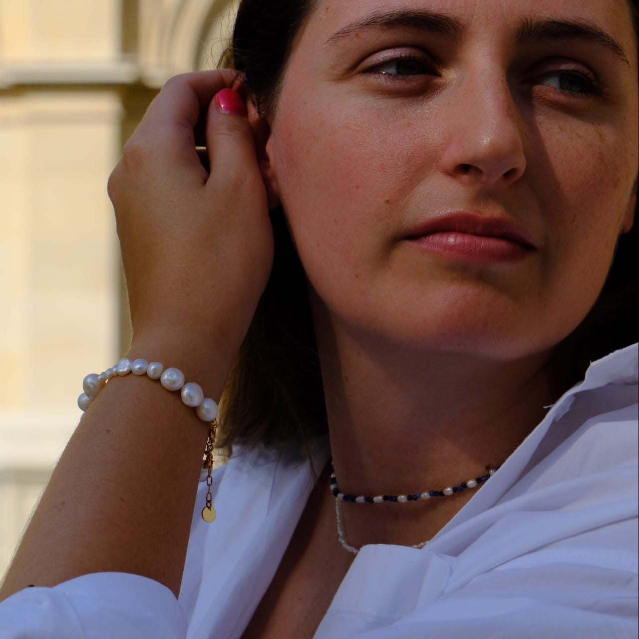 A close-up lifestyle shot of a woman wearing a white pearl beaded bracelet with a small gold charm, paired with a black and white beaded necklace, set against a blurred background of a classical building.