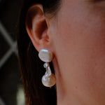 A close-up of a person's ear wearing a double-drop pearl earring with a small silver accent, paired with a matching pearl necklace.