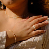 A close-up lifestyle shot of a woman wearing a matching jewelry set, featuring a delicate gold chain necklace, a gold beaded bracelet, and a gold beaded ring on her finger.