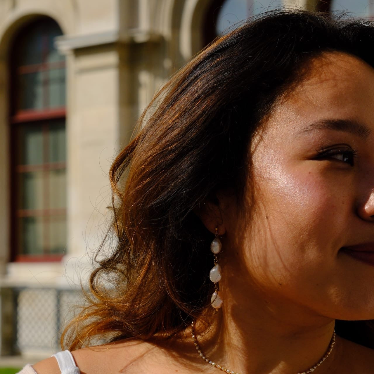 A close-up lifestyle shot of a woman with dark brown hair wearing a delicate gold chain necklace and matching pearl drop earrings, set against a blurred background of a classical stone building.