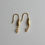 A pair of gold dangle earrings with teardrop-shaped metal drops and small white pearl beads hanging below, displayed on a light gray textured background.