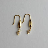 A pair of gold dangle earrings with teardrop-shaped metal drops and small white pearl beads hanging below, displayed on a light gray textured background.