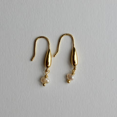 A pair of gold dangle earrings with teardrop-shaped metal drops and small white pearl beads hanging below, displayed on a light gray textured background.