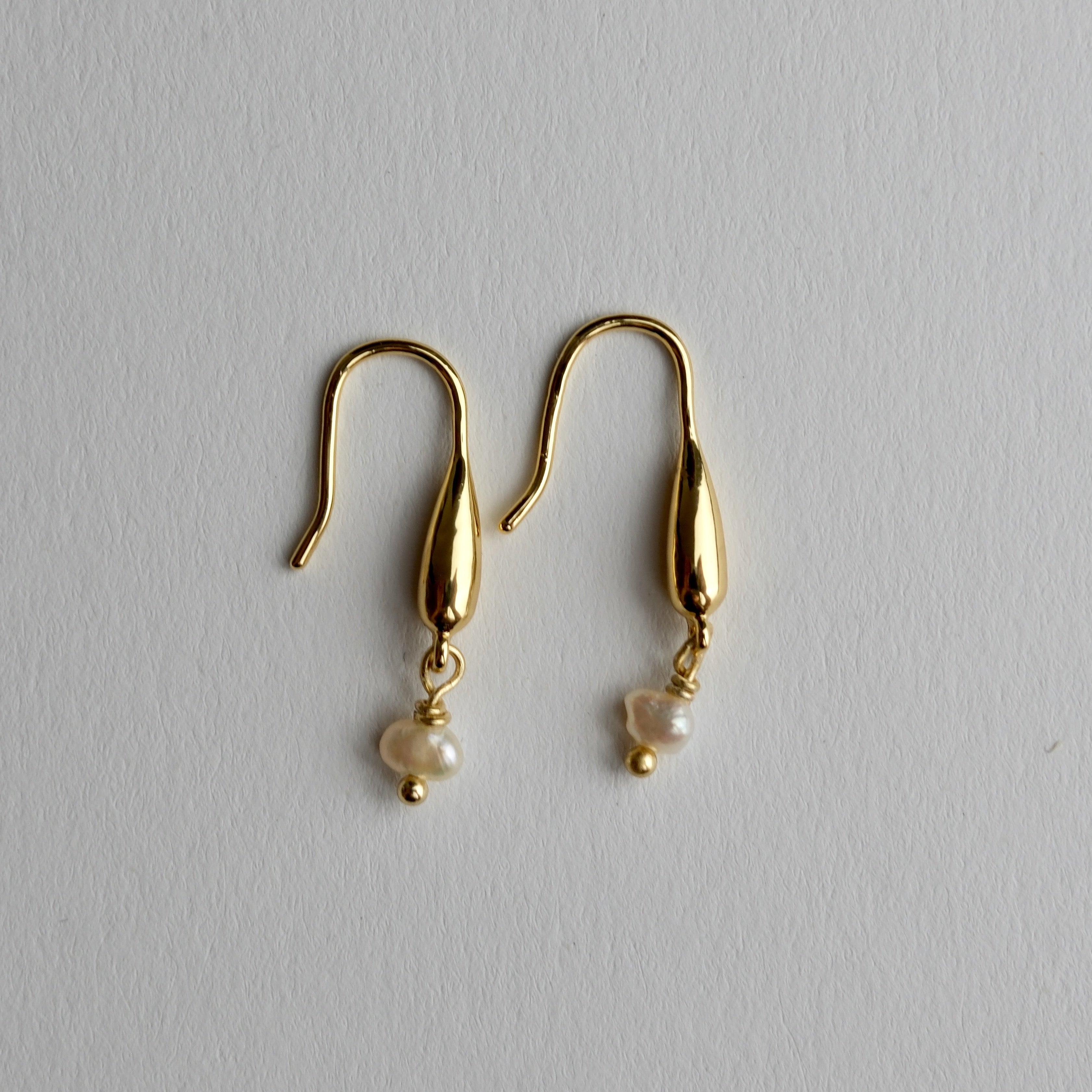 A pair of gold dangle earrings with teardrop-shaped metal drops and small white pearl beads hanging below, displayed on a light gray textured background.