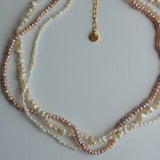 A multi-strand beaded necklace featuring white and pinkish-beige baroque pearls with a gold-tone chain and a small round gold tag, displayed on a light gray background.