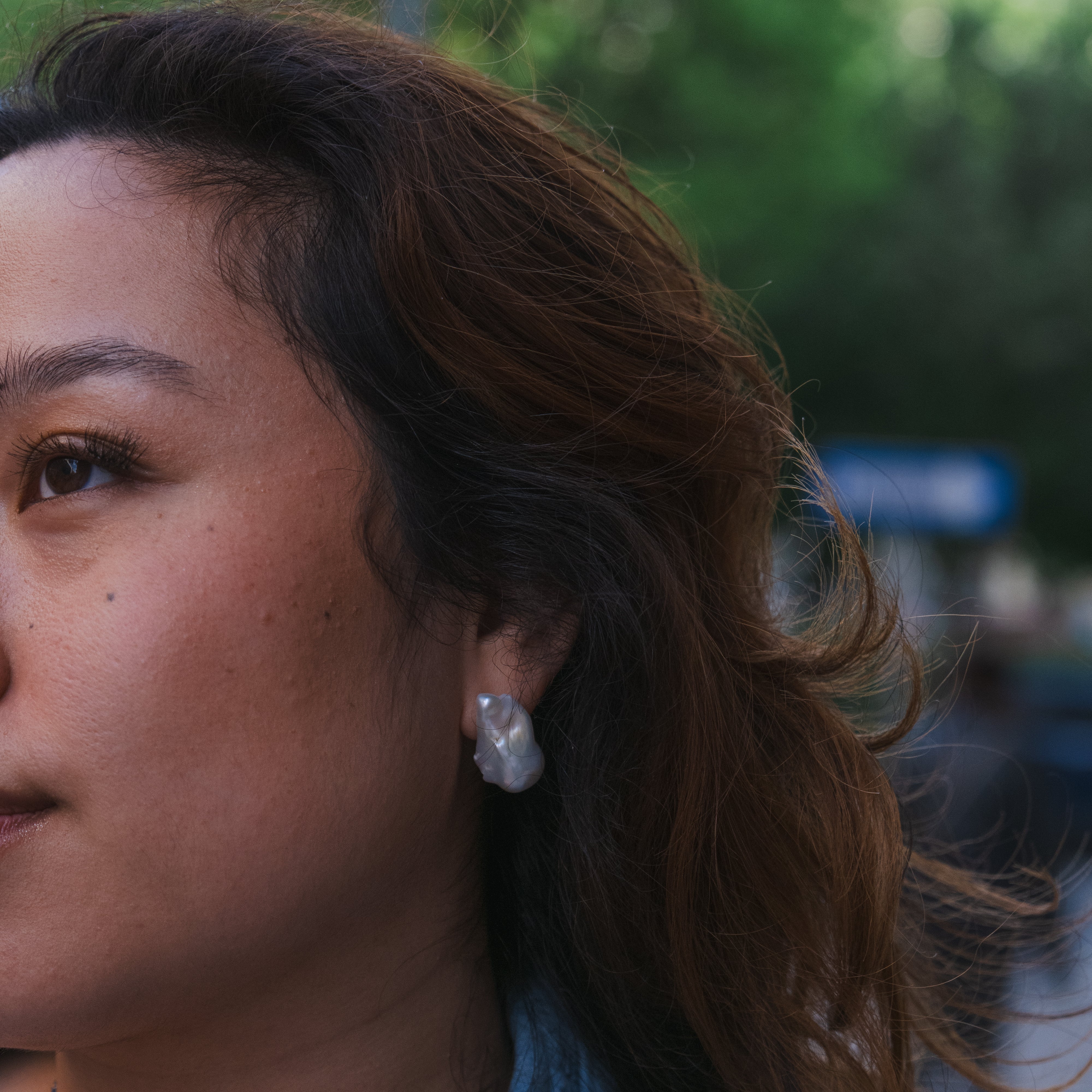 A close-up side profile of a woman with brown hair wearing a textured white pearl earring and a dark beaded necklace, wearing a blue denim shirt against a blurred outdoor background.