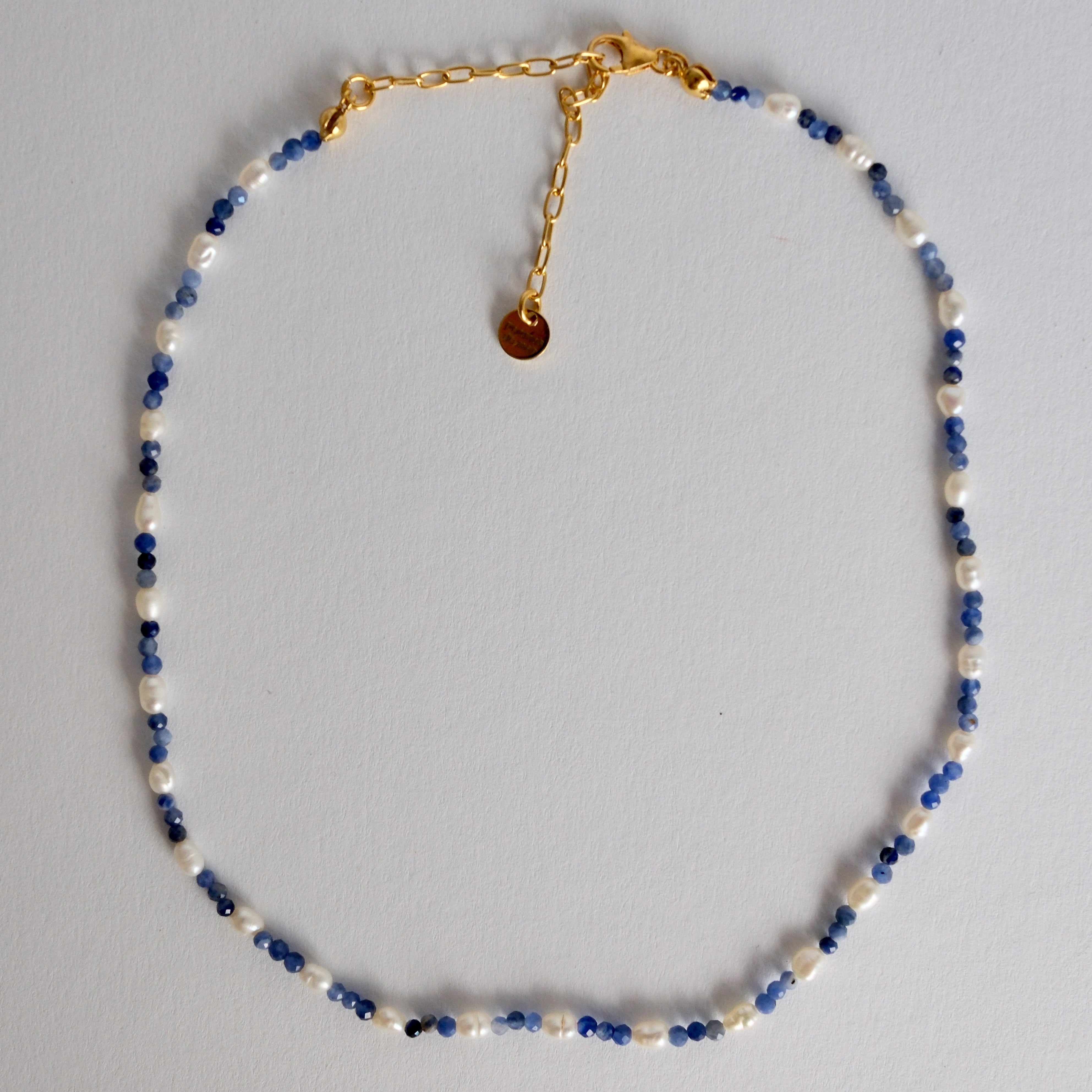 A delicate beaded necklace with alternating white pearl-like beads and small blue faceted beads, finished with a gold-tone chain extender and a small round gold disc charm, laid flat on a light gray background.