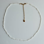 A delicate beaded necklace made of small white beads with a gold-tone chain extender and a small round gold disc charm, laid flat on a light gray background.