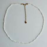A delicate beaded necklace made of small white beads with a gold-tone chain extender and a small round gold disc charm, laid flat on a light gray background.
