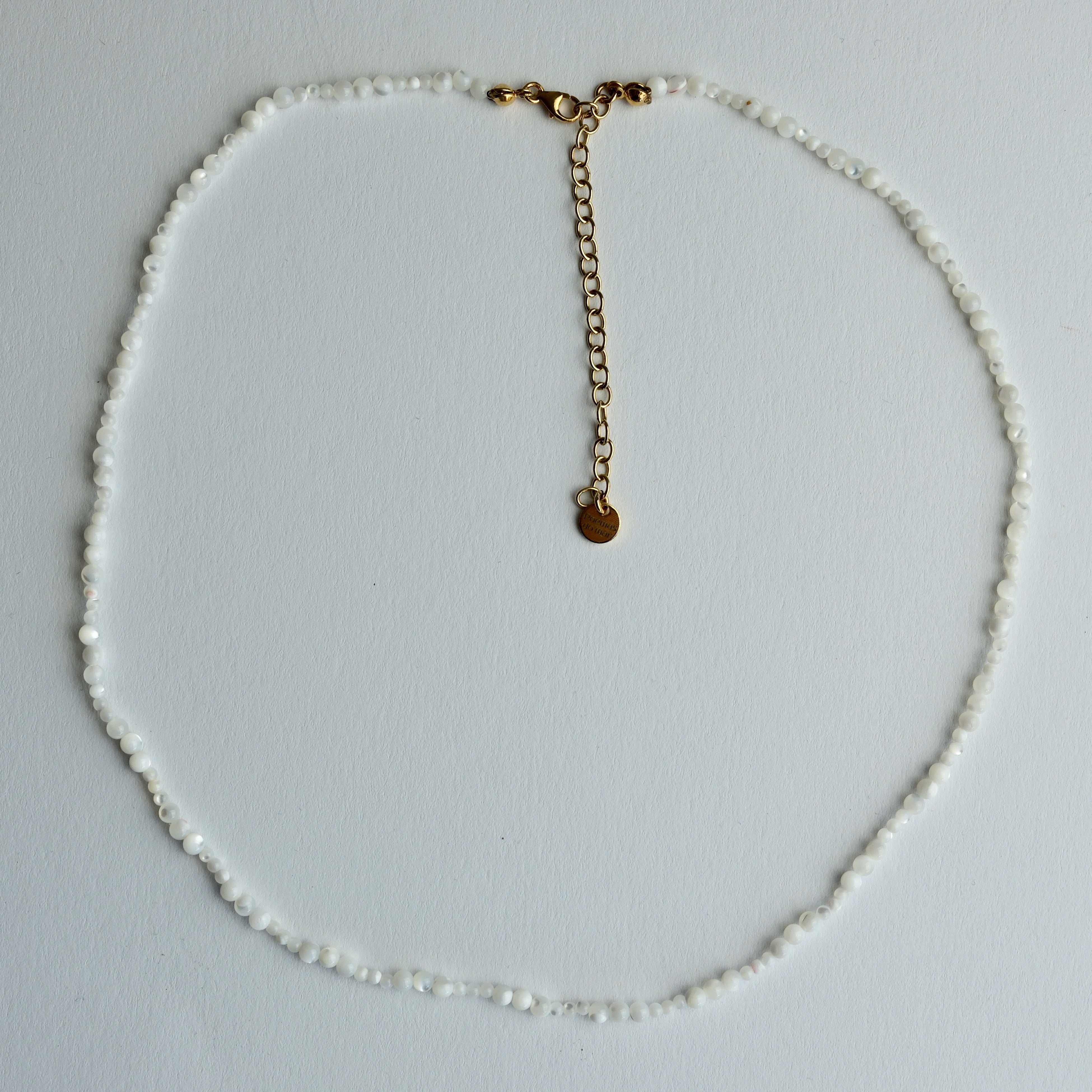 A delicate beaded necklace made of small white beads with a gold-tone chain extender and a small round gold disc charm, laid flat on a light gray background.