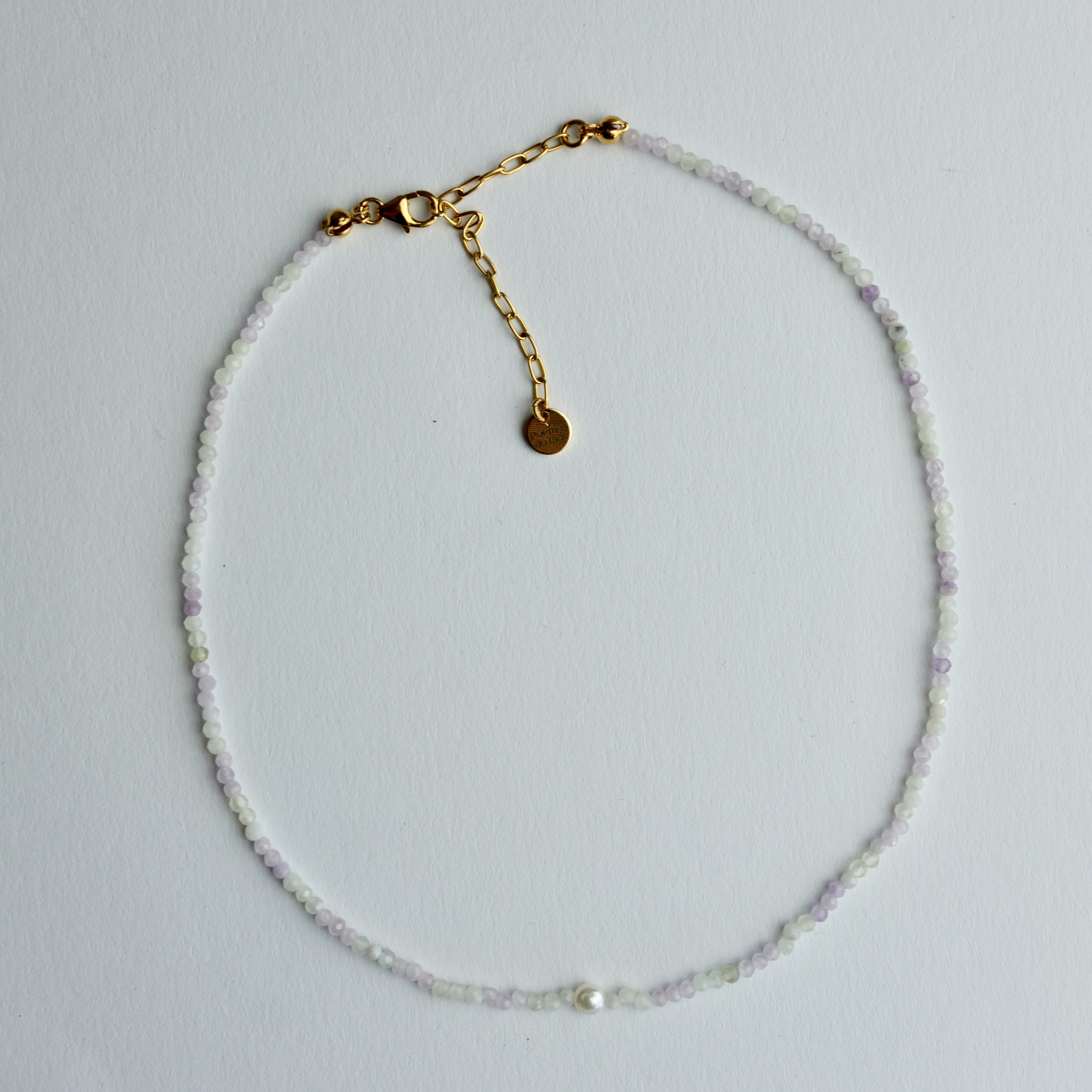 A delicate beaded necklace with iridescent white beads, subtle lavender accents, and a gold-tone clasp with a heart charm and extender chain, displayed on a light gray background.