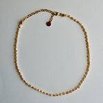 A delicate gold chain necklace with a small golden beads laid out in a circle on a light gray background.