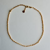 A delicate gold chain necklace with a small golden beads laid out in a circle on a light gray background.