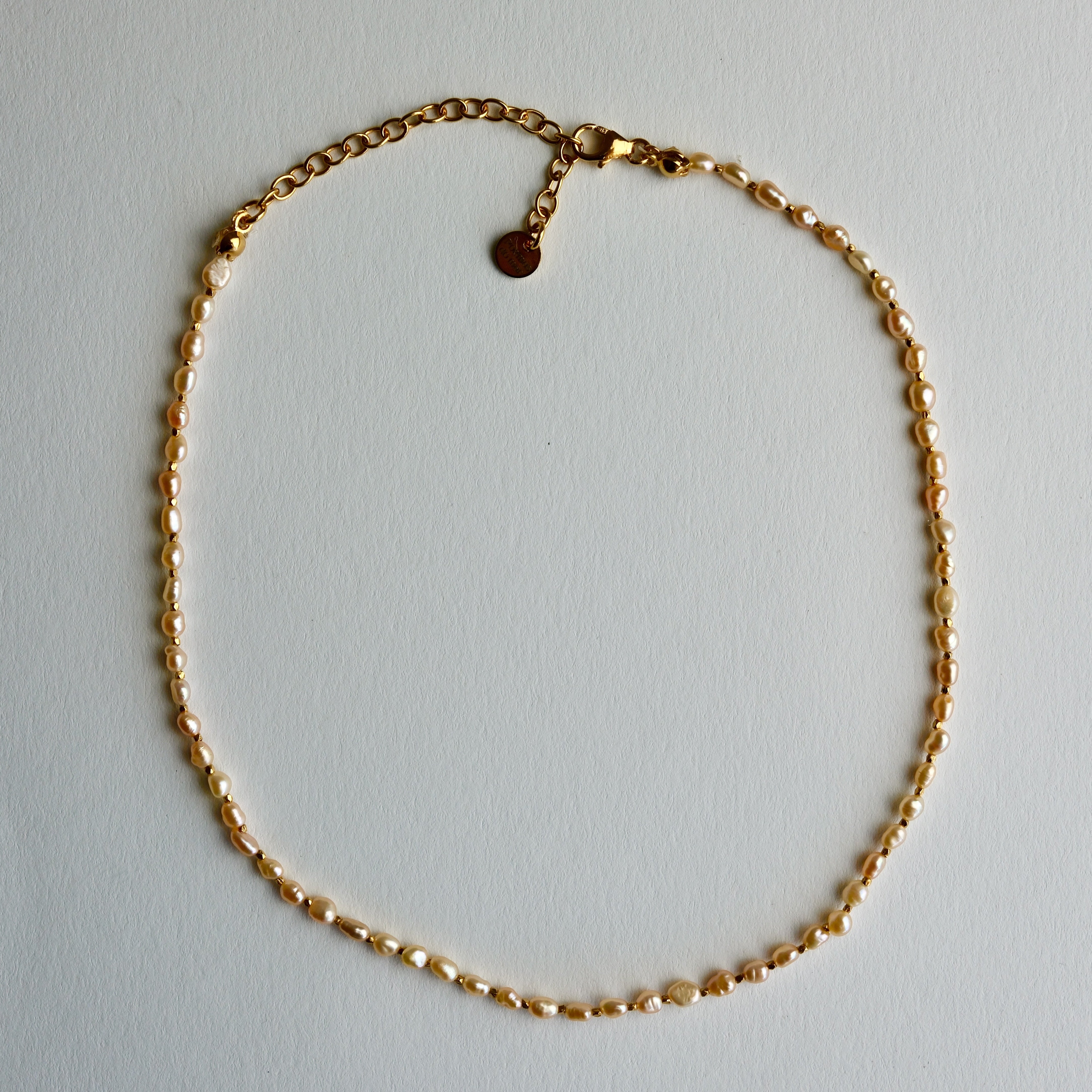 A delicate gold chain necklace with a small golden beads laid out in a circle on a light gray background.