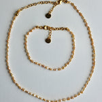 A gold chain necklace and matching bracelet displayed on a light gray background. The jewelry features small, irregular pearl-like beads woven throughout the chain, with gold lobster clasp closures and circular brand tags.