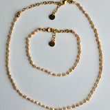 A gold chain necklace and matching bracelet displayed on a light gray background. The jewelry features small, irregular pearl-like beads woven throughout the chain, with gold lobster clasp closures and circular brand tags.