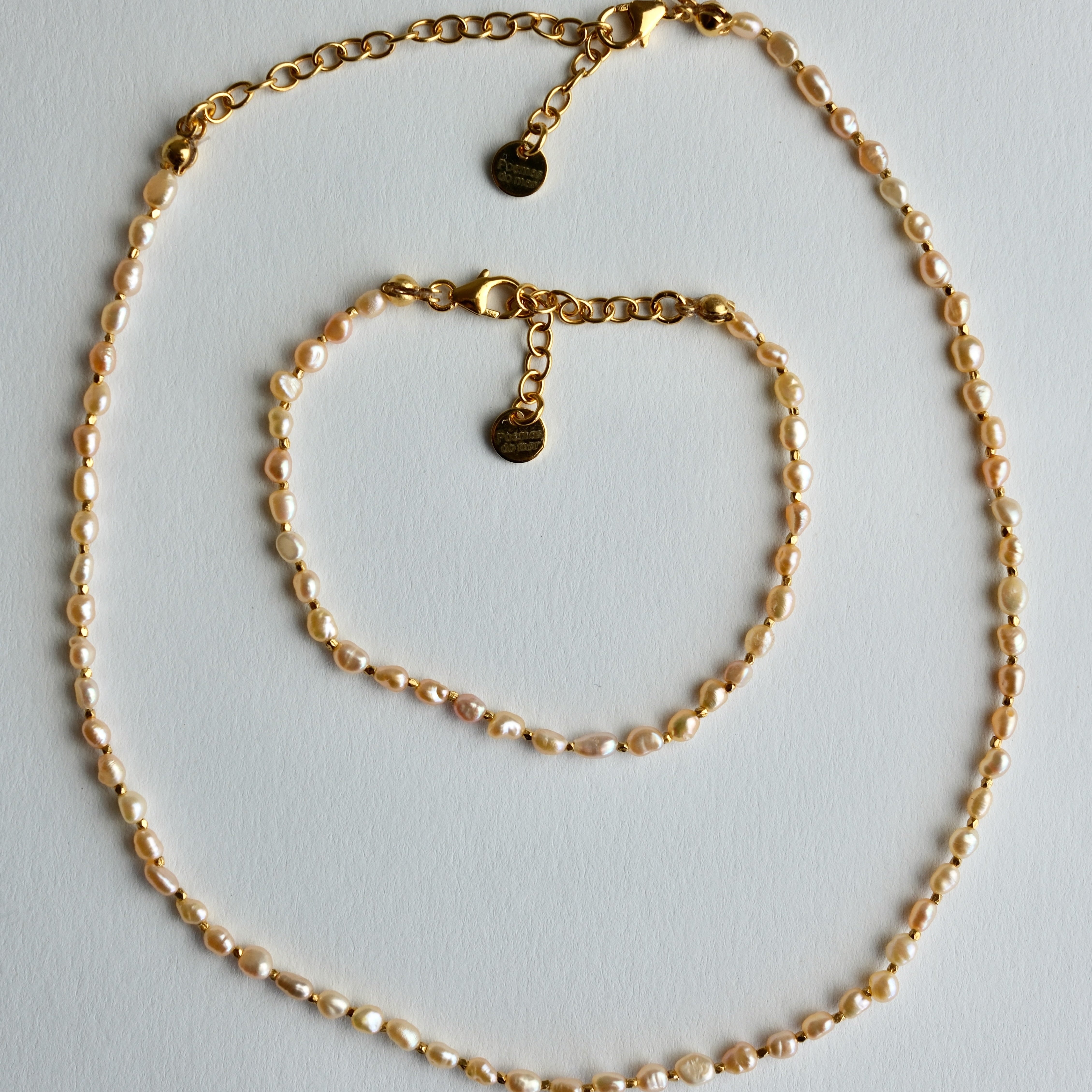 A gold chain necklace and matching bracelet displayed on a light gray background. The jewelry features small, irregular pearl-like beads woven throughout the chain, with gold lobster clasp closures and circular brand tags.