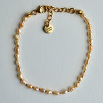 A delicate gold chain bracelet featuring irregular, baroque-style pearl beads in a soft champagne pink color, complete with a lobster clasp and a heart-shaped charm engraved with 'poemas do mar'.