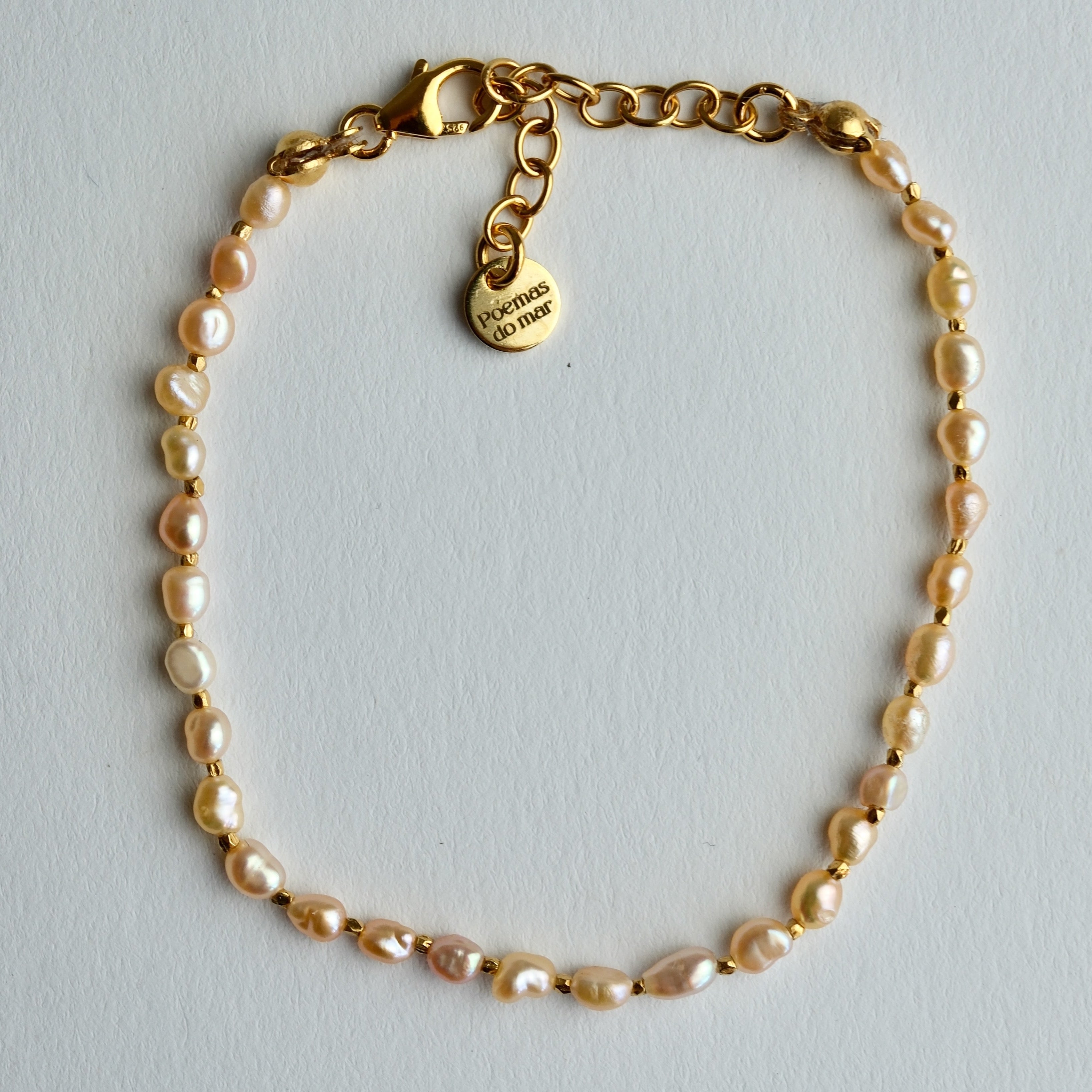A delicate gold chain bracelet featuring irregular, baroque-style pearl beads in a soft champagne pink color, complete with a lobster clasp and a heart-shaped charm engraved with 'poemas do mar'.