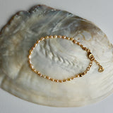 A delicate gold chain bracelet with small pearl-like beads and a lobster clasp, displayed on a large iridescent shell against a light gray background.