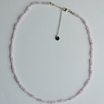 A delicate beaded necklace made of small light pink faceted beads with a silver-tone chain extender and a small round black charm, laid flat on a white background.