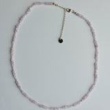 A delicate beaded necklace made of small light pink faceted beads with a silver-tone chain extender and a small round black charm, laid flat on a white background.