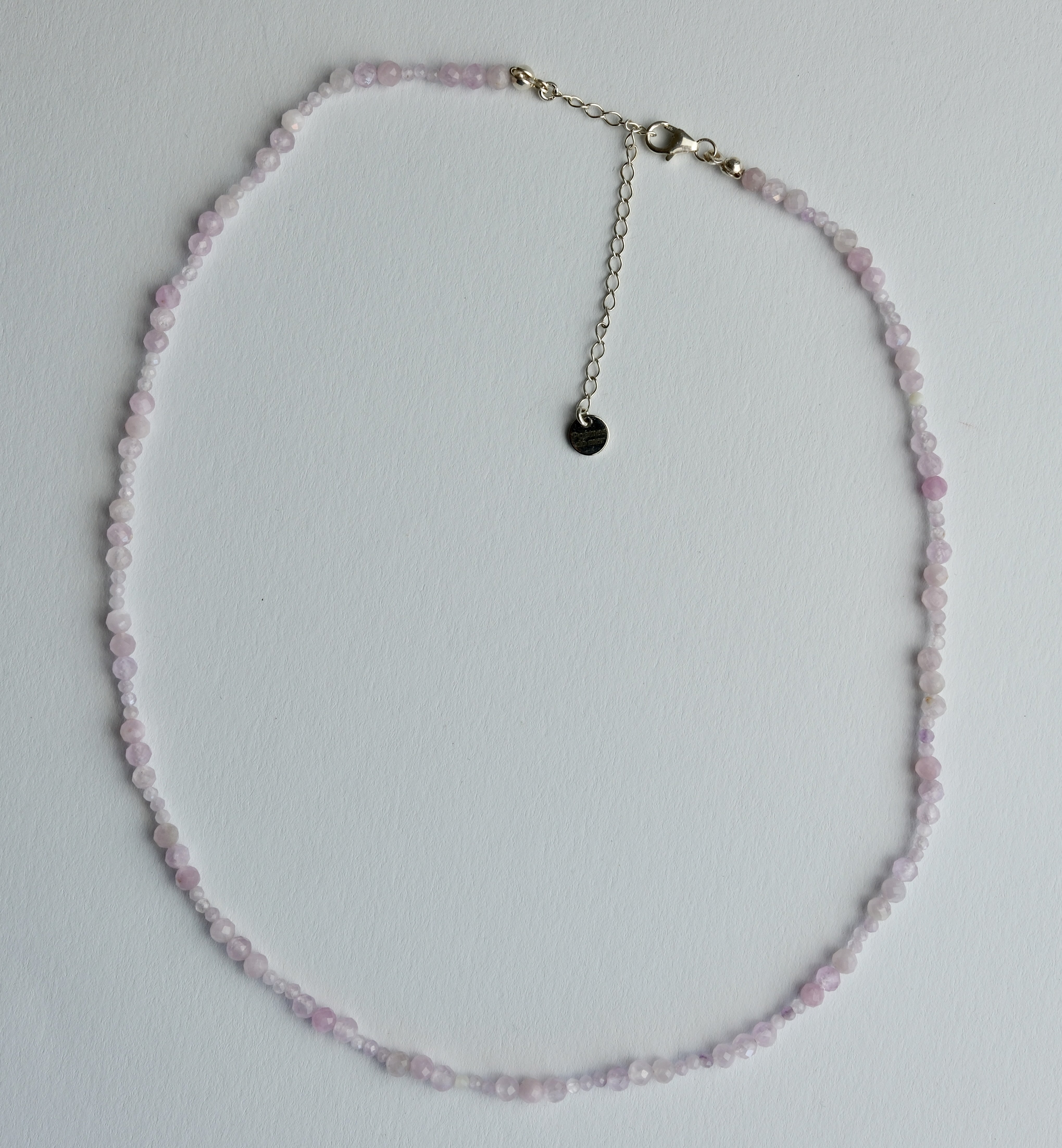 A delicate beaded necklace made of small light pink faceted beads with a silver-tone chain extender and a small round black charm, laid flat on a white background.