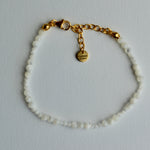 A delicate beaded bracelet with small white beads and a gold chain clasp, featuring a small gold tag engraved with the brand's logo, 