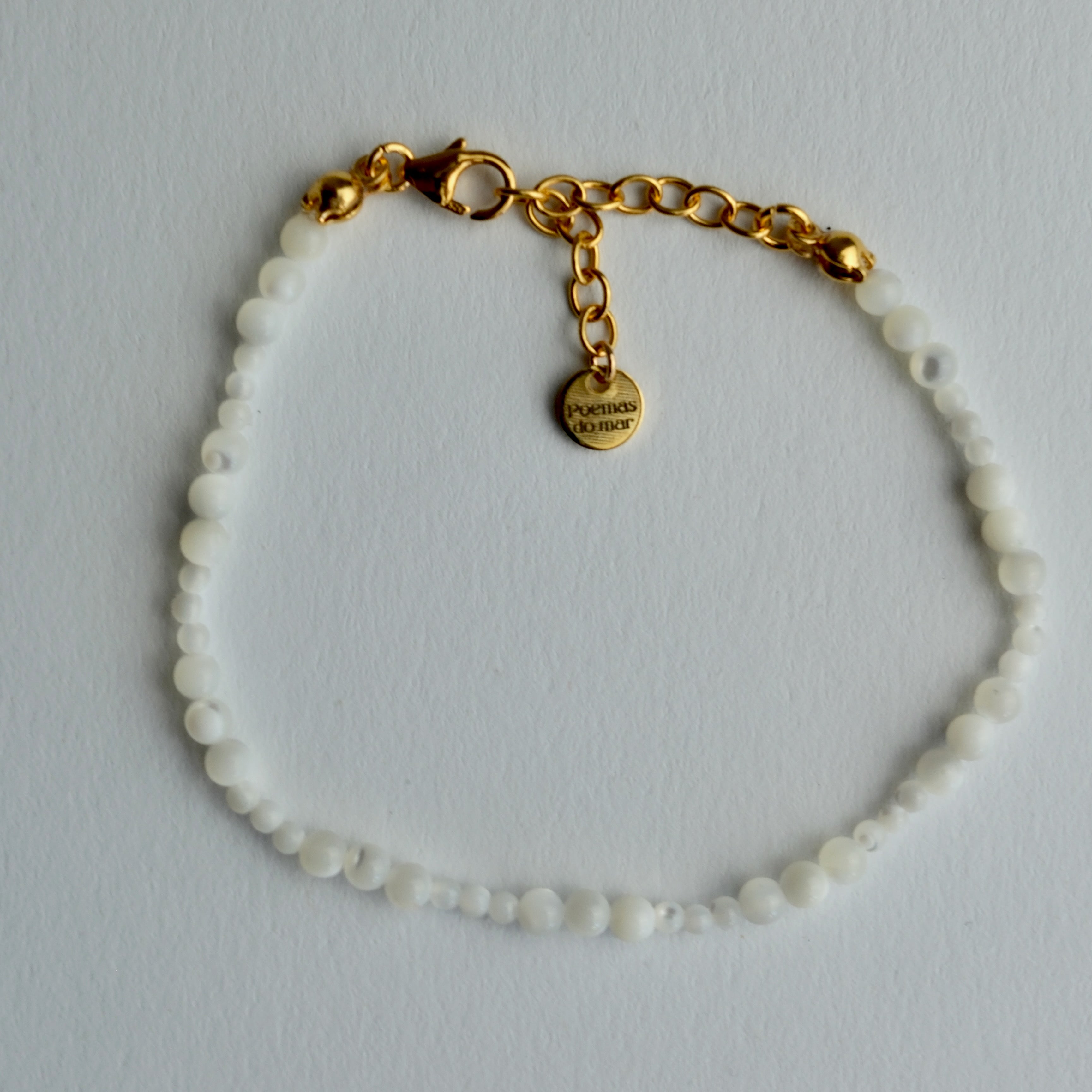 A delicate beaded bracelet with small white beads and a gold chain clasp, featuring a small gold tag engraved with the brand's logo, 