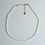 A delicate gold chain necklace with small, irregular cream-colored beads, arranged in a circle on a light gray background. It features a lobster clasp closure and an adjustable gold chain extender with a small round tag.