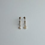 A pair of small gold huggie hoop earrings with dangling white freshwater pearls, shown on a plain light gray background.