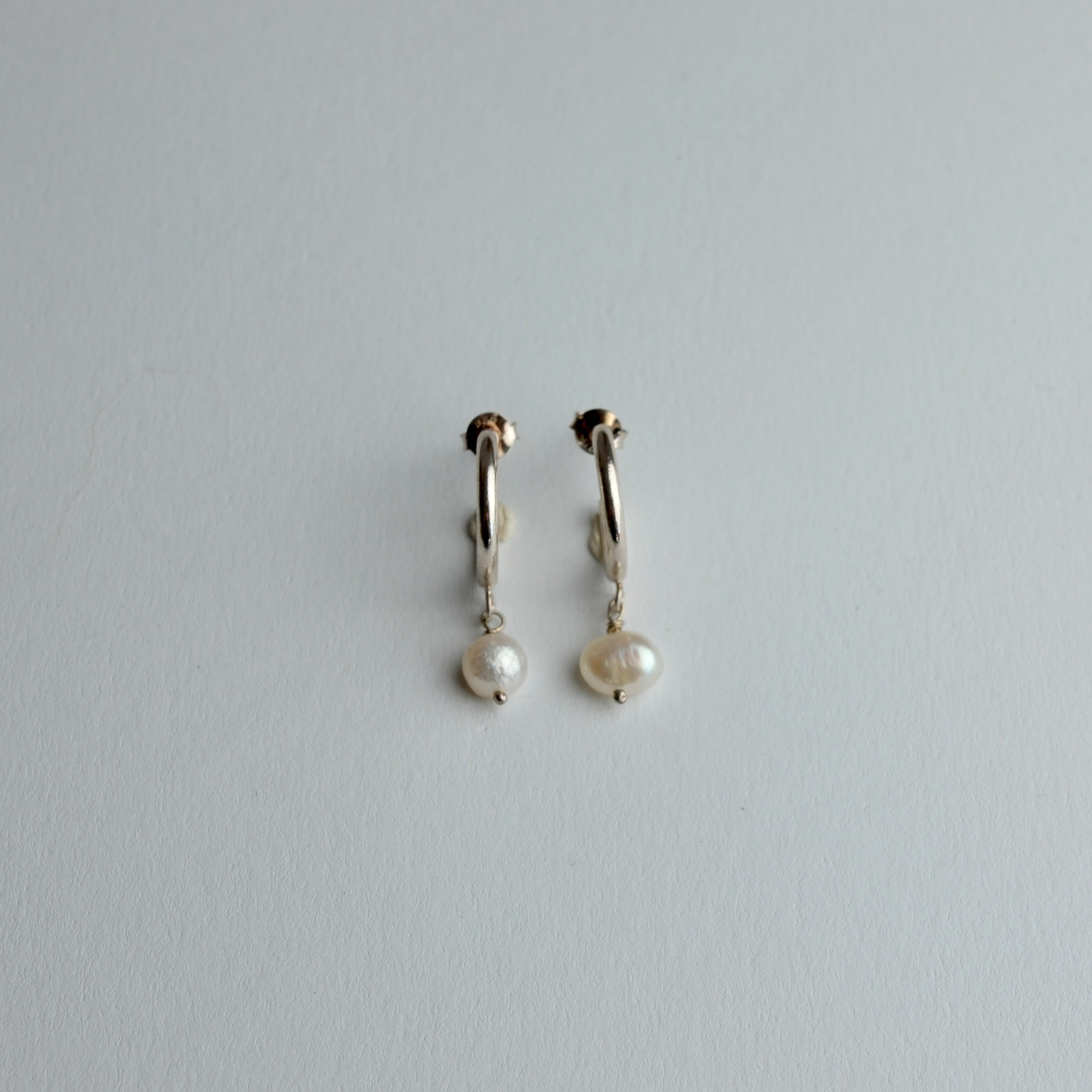 A pair of small gold huggie hoop earrings with dangling white freshwater pearls, shown on a plain light gray background.