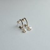 A pair of gold huggie hoop earrings with dangling baroque pearl drops, photographed on a clean, light gray textured surface.