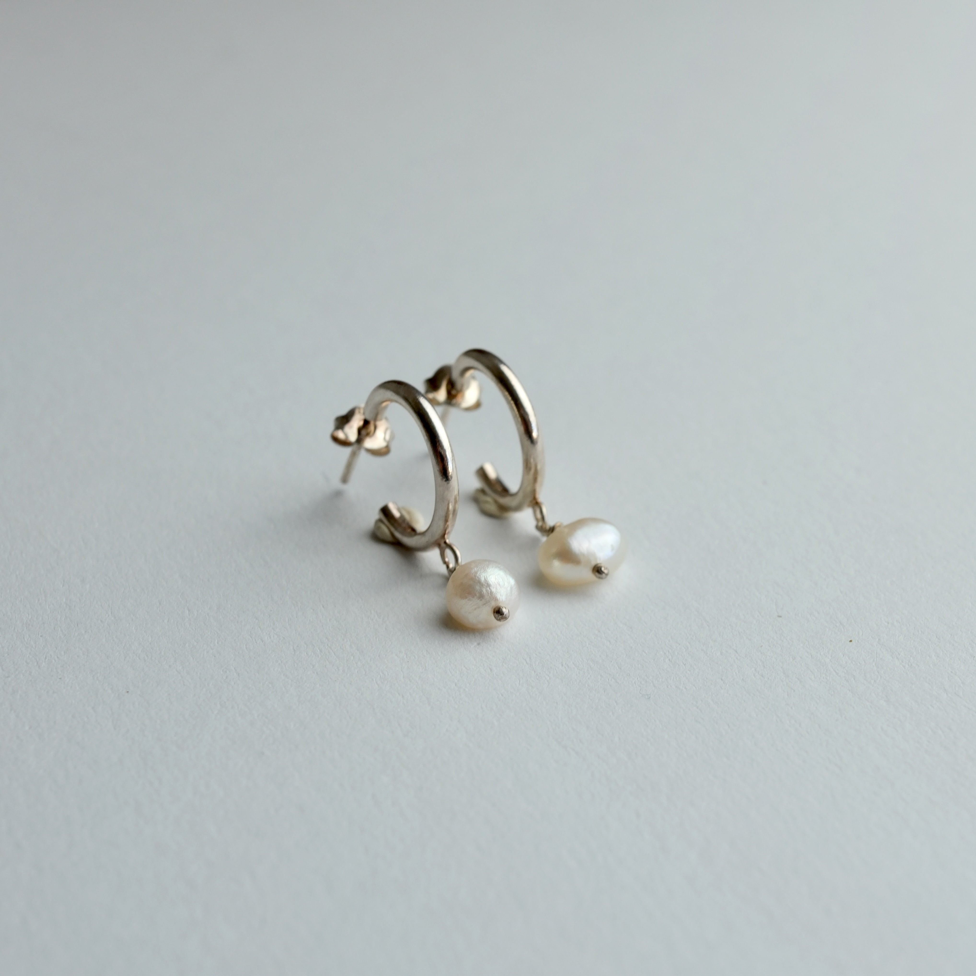 A pair of gold huggie hoop earrings with dangling baroque pearl drops, photographed on a clean, light gray textured surface.