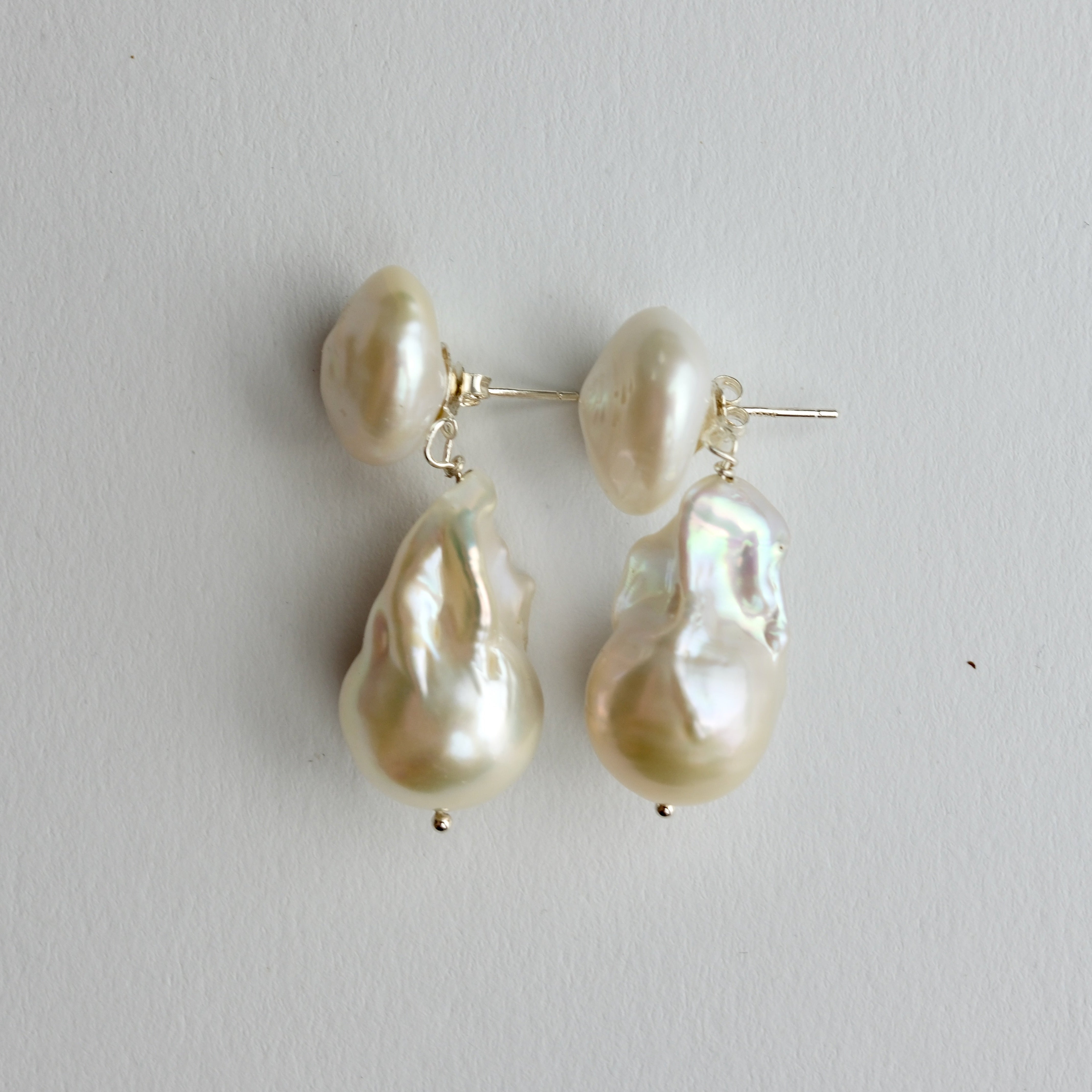 A pair of baroque pearl drop earrings with silver post settings, displayed on a neutral light gray background.