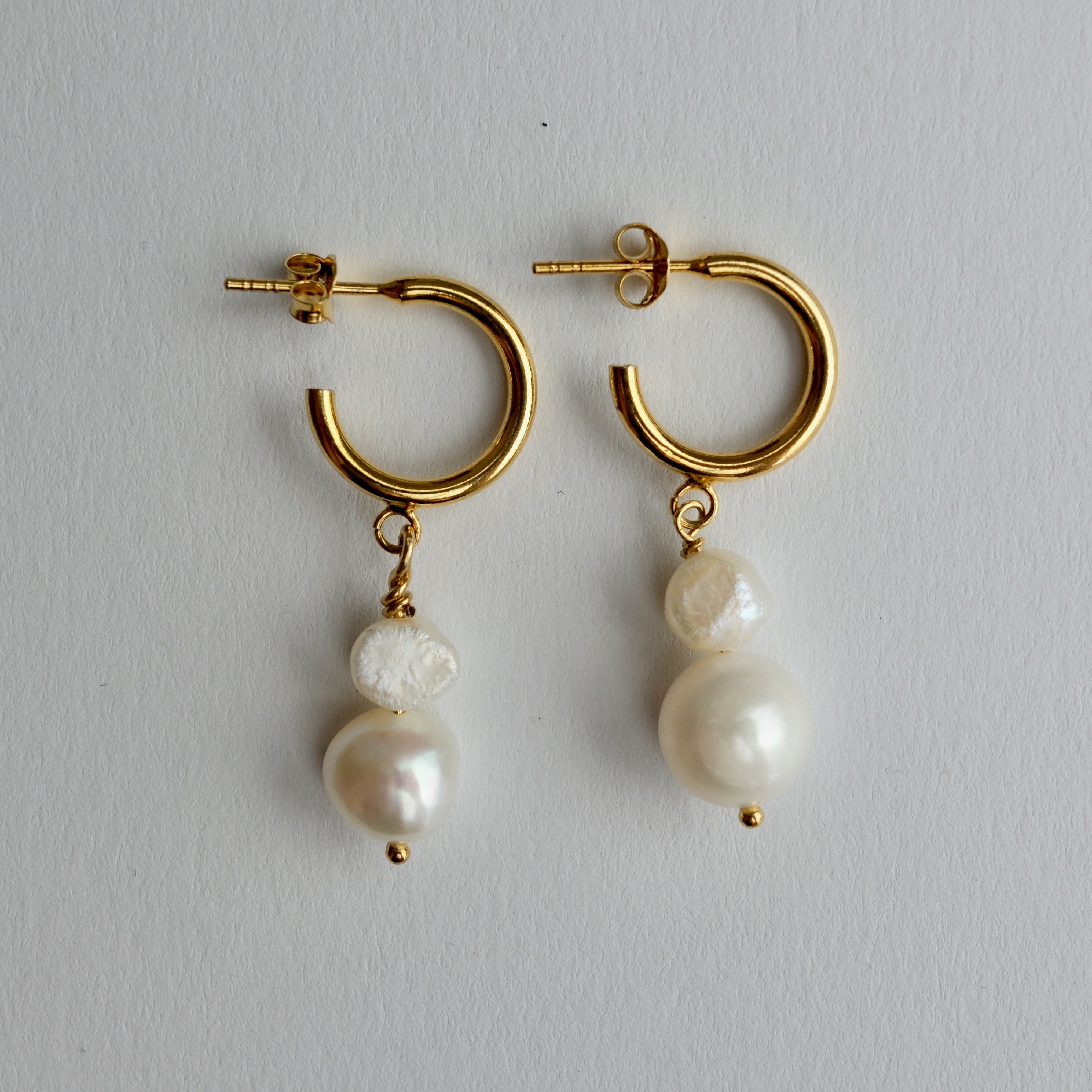 A pair of gold hoop earrings with dangling baroque pearl beads, displayed on a neutral light gray background.