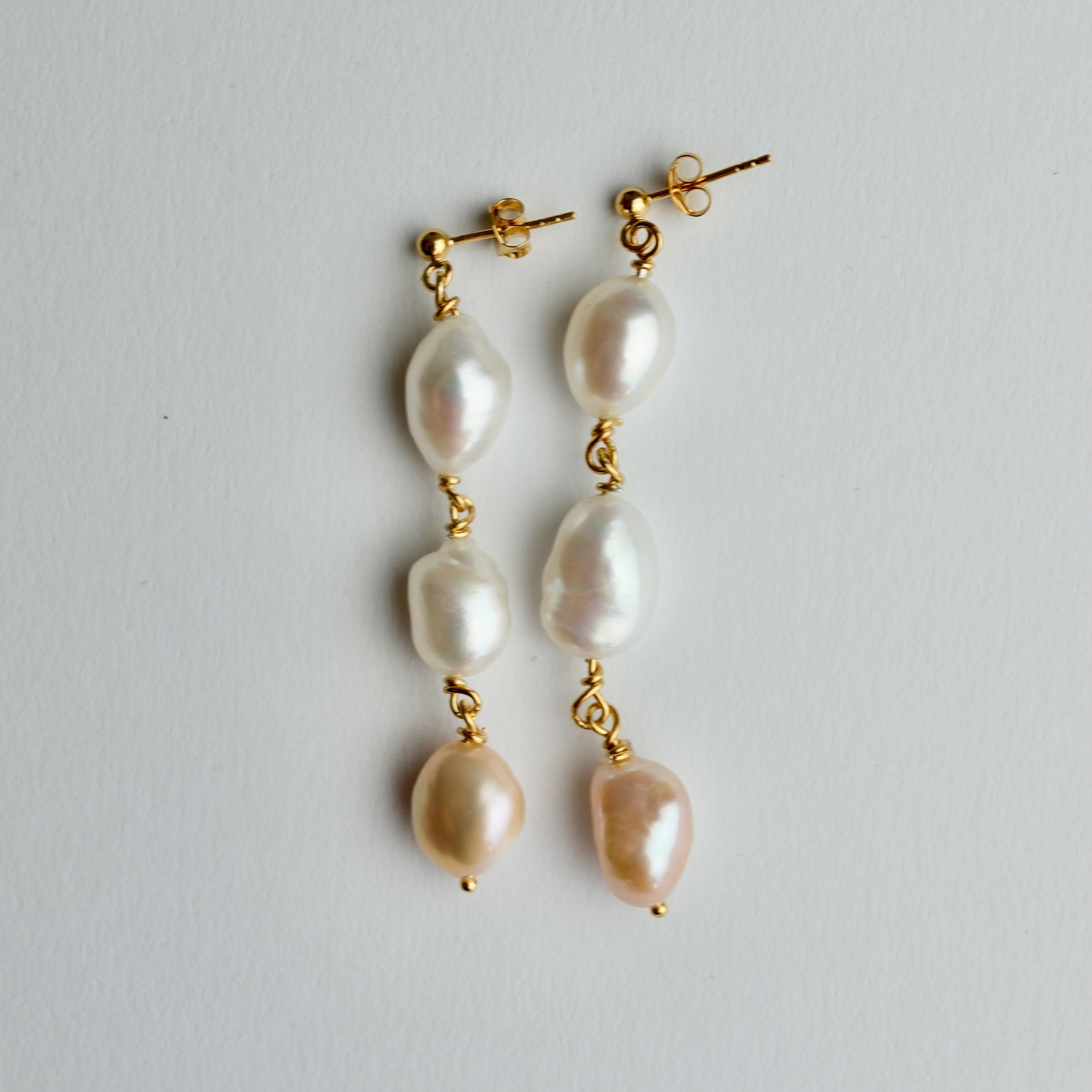 A pair of gold drop earrings featuring three irregular, lustrous white baroque pearls suspended vertically on each earring, shown against a plain light gray background.