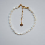 A delicate bracelet made of white mother-of-pearl oval beads with a gold chain extender and lobster clasp, featuring a small round gold tag, displayed on a light gray background.