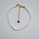 A delicate bracelet made of white mother-of-pearl oval beads with a gold chain extender and lobster clasp, featuring a small round gold tag, displayed on a light gray background.