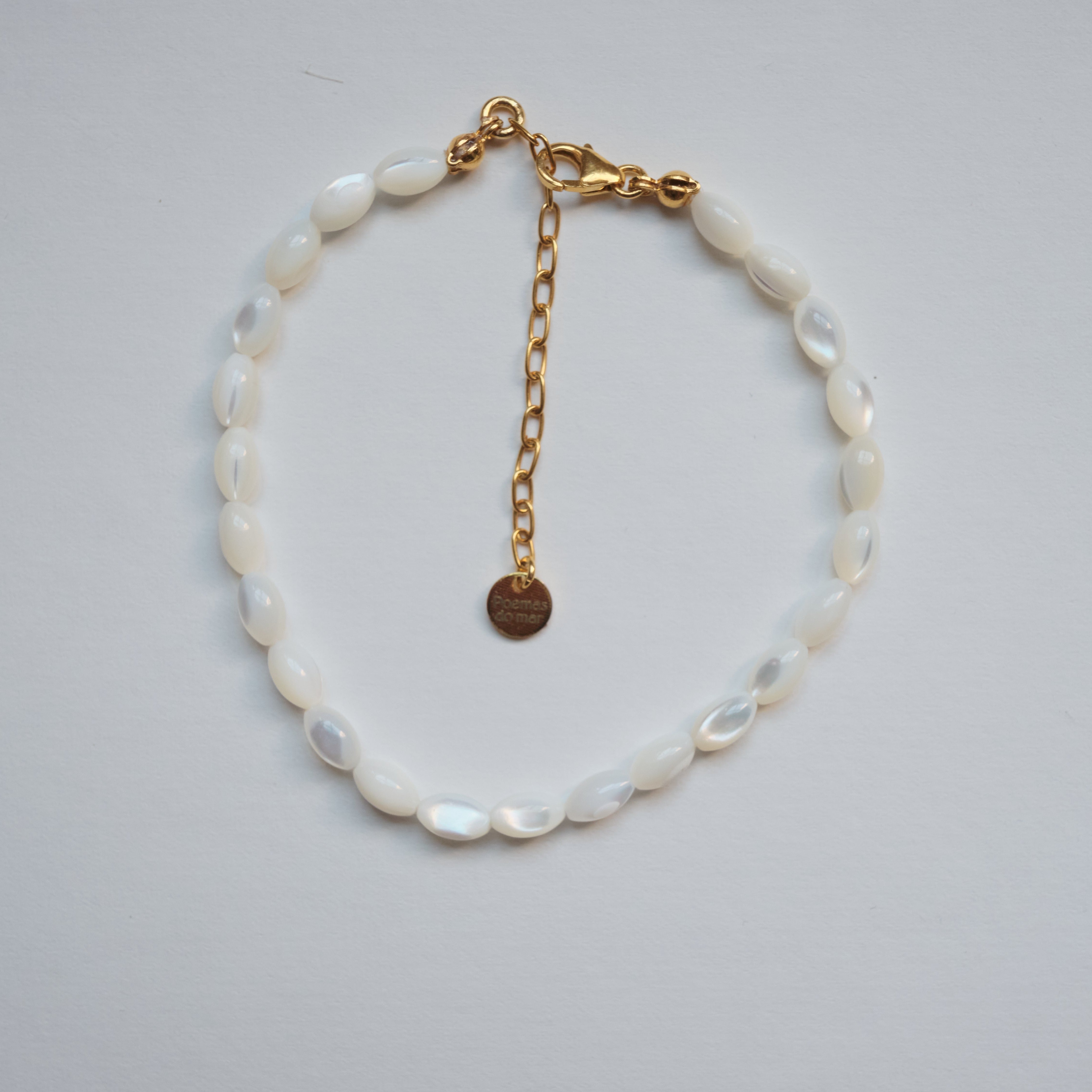 A delicate bracelet made of white mother-of-pearl oval beads with a gold chain extender and lobster clasp, featuring a small round gold tag, displayed on a light gray background.