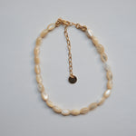 A beaded necklace made of irregular cream-colored shell-like beads with a gold-tone chain extender and small round tag, photographed on a plain light gray background.