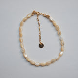A beaded necklace made of irregular cream-colored shell-like beads with a gold-tone chain extender and small round tag, photographed on a plain light gray background.